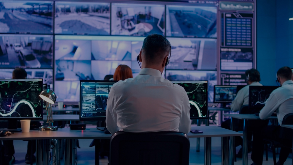 Back view of young man with headset watching surveillance footage and map on computers and speaking with female colleague while working in intelligence agency